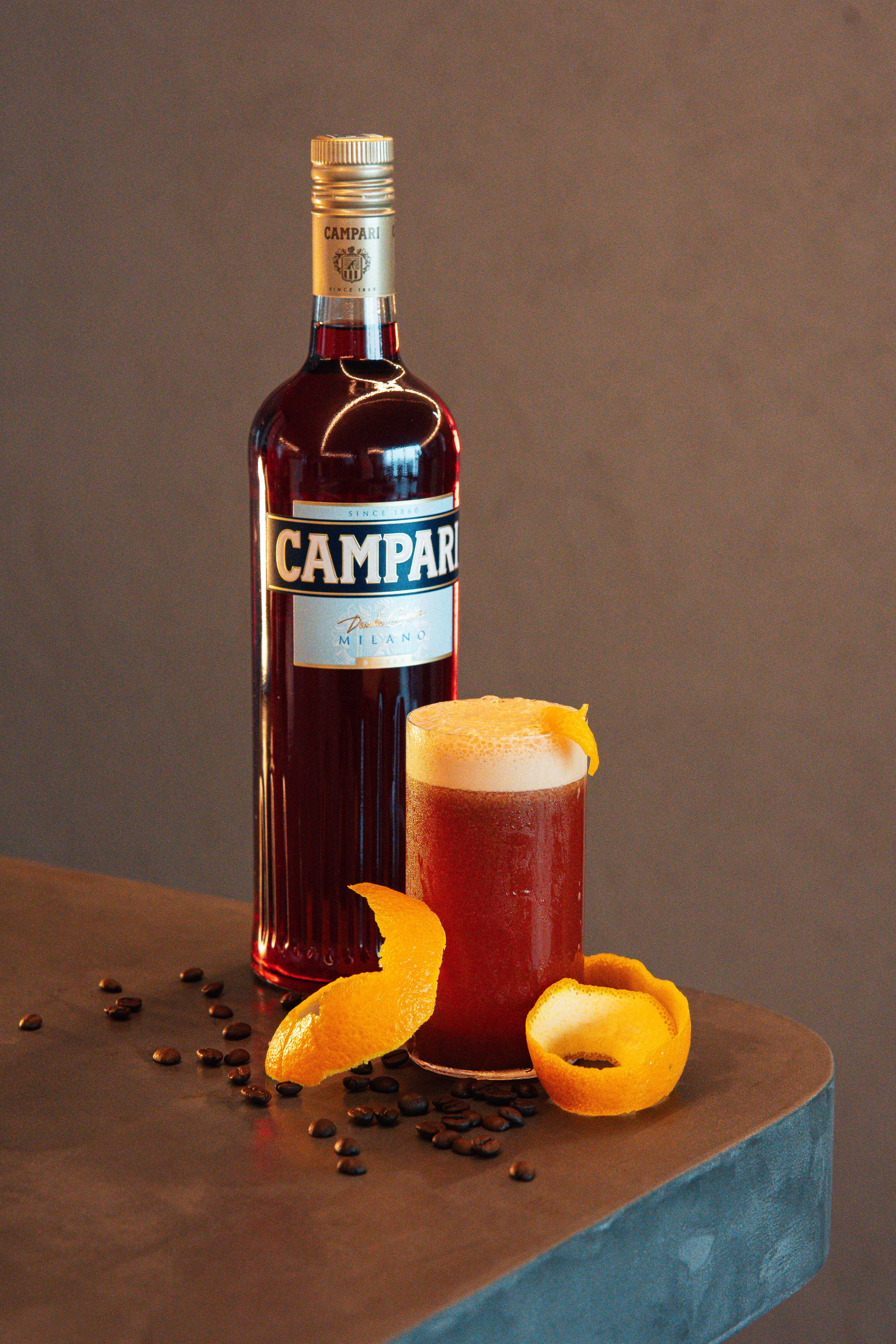 A Campari bottle beside a frothy orange Garibaldi with a citrus twist